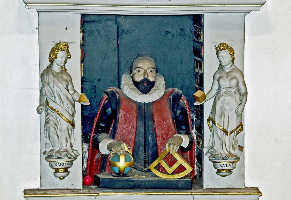 Wall monument: bearded man in lay costume holding mathematical instruments, set in a book-lined tabernacle, surrounded by allegorical figures that carry geometrical symbols. Wall monument: bearded man in lay costume holding mathematical instruments, set in a book-lined tabernacle, surrounded by allegorical figures that carry geometrical symbols.