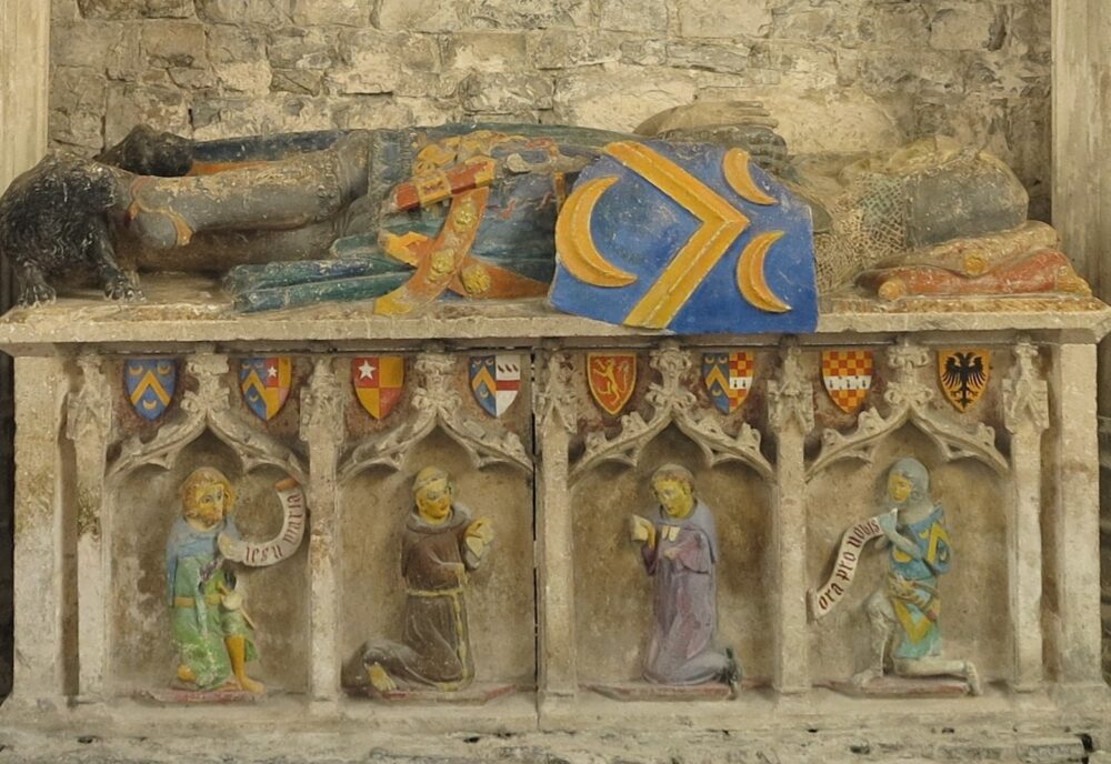 Tomb chest with effigy of mailed knight. On the chest are kneeling figures of civilian, a monk, a secular priest and a knight. The monk and priest have beeoks, the others have speech scrolls but the wording on the scrolls is modern.