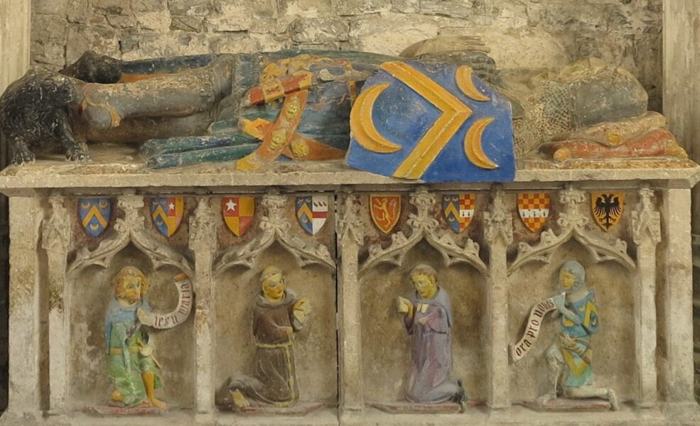 Tomb chest with effigy of mailed knight. On the chest are kneeling figures of civilian, a monk, a secular priest and a knight. The monk and priest have beeoks, the others have speech scrolls but the wording on the scrolls is modern.