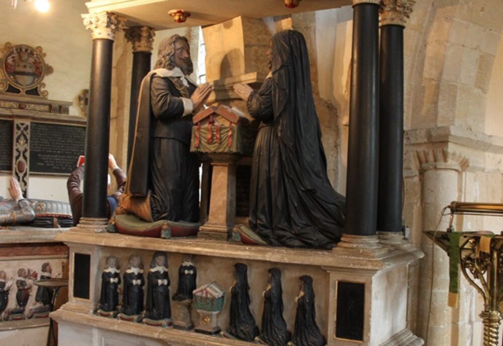 Tomb carving against arcade pillar. Pediment with heraldic shield. Couple kneeling either side of prayer desk. Miniature children kneeling underneath.