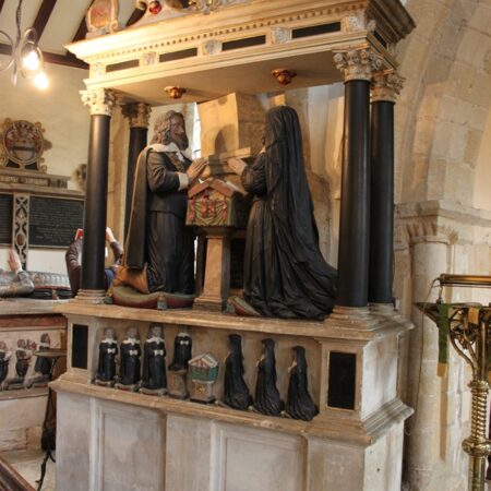 Tomb carving against arcade pillar. Pediment with heraldic shield. Couple kneeling either side of prayer desk. Miniature children kneeling underneath.