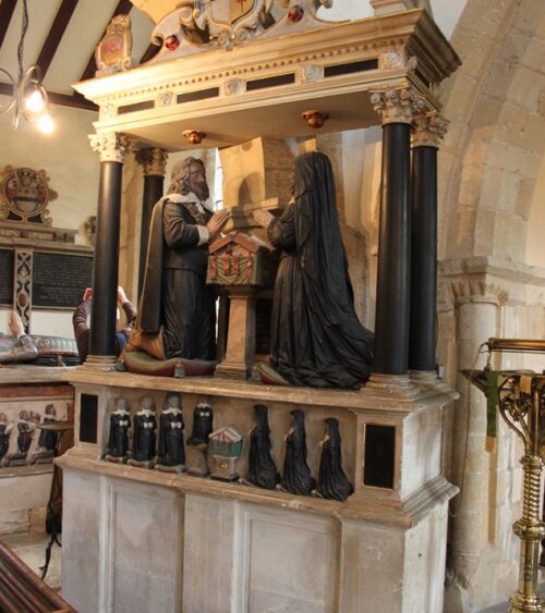 Tomb carving against arcade pillar. Pediment with heraldic shield. Couple kneeling either side of prayer desk. Miniature children kneeling underneath.