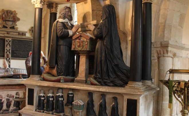 Tomb carving against arcade pillar. Pediment with heraldic shield. Couple kneeling either side of prayer desk. Miniature children kneeling underneath.