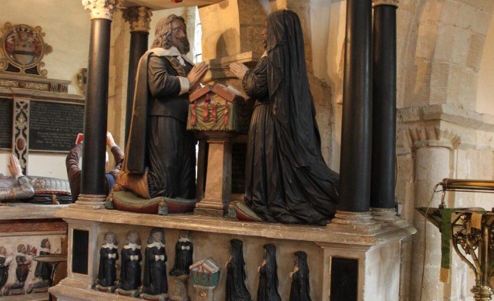 Tomb carving against arcade pillar. Pediment with heraldic shield. Couple kneeling either side of prayer desk. Miniature children kneeling underneath.