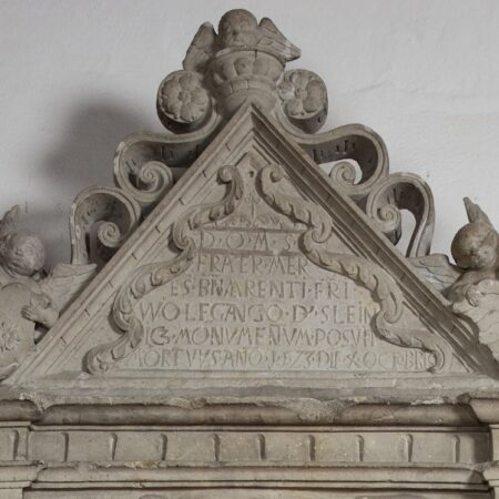 Tympanum above the cadaver effigy with floral decoration, cherubs and inscription D(EO) . O(PTIMO) . M(AXIMO) . S(ACRVM). FRATER . M(A)ER E(N)S . B(E)NEMERENTI . F(RAT)RI . WOLFGANGO . D(OMINO) . SLEIN ICZ . MONVMENTVM . POSVIT MORTVVS . AN(N)O . 1523 . X . OCTOBRIS