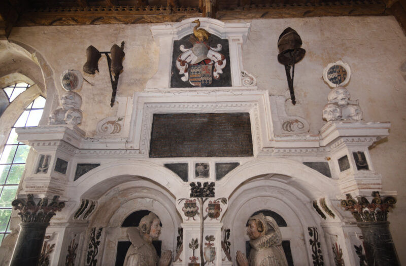Upper part of wall monument: heads of kneeling wives, heraldic decoration, gauntlets and helm