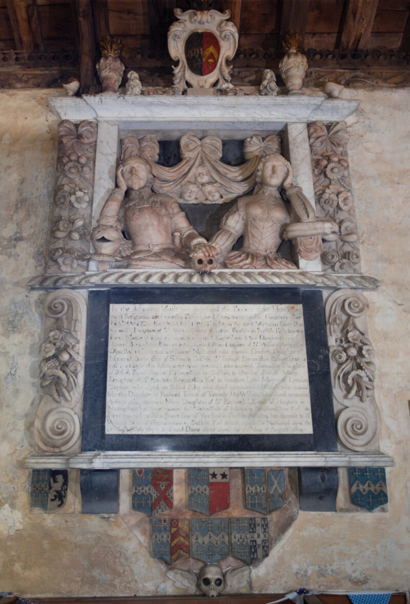 Wall monument: busts of male and female in mid C17 costume, hands on a skull. Surrounded by ornate pillars, swags of fruit and foliage and cherubs.