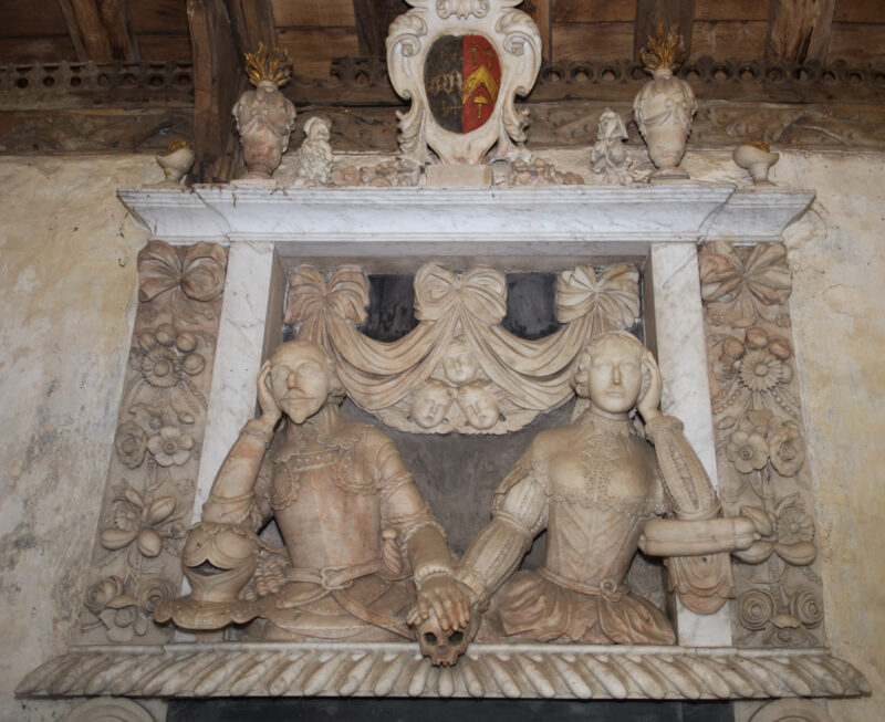 Detail of wall monument with busts of male and female in mid C17 costume, hands on skull.