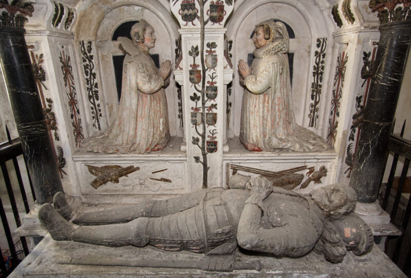 Recumbent effigy of man in armour, 2 kneeling female figures behind. Decorated with scrolls, heraldry and armaments.