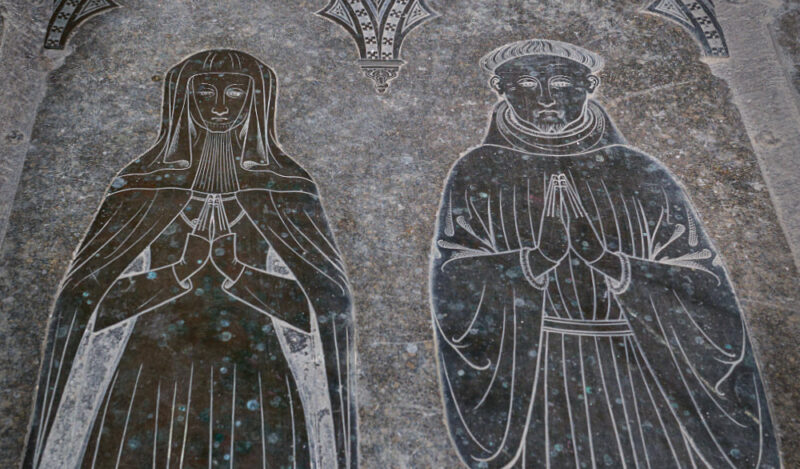 Monumental brasses of couple. She wears veil, barbe and mantle, he wears long gown.