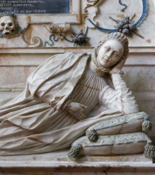 Tomb carving. Young woman in early C17 costume reclining on cushions in niche with baroque decoration and a skull
