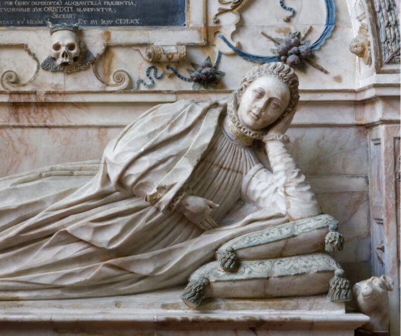 Tomb carving. Young woman in early C17 costume reclining on cushions in niche with baroque decoration and a skull