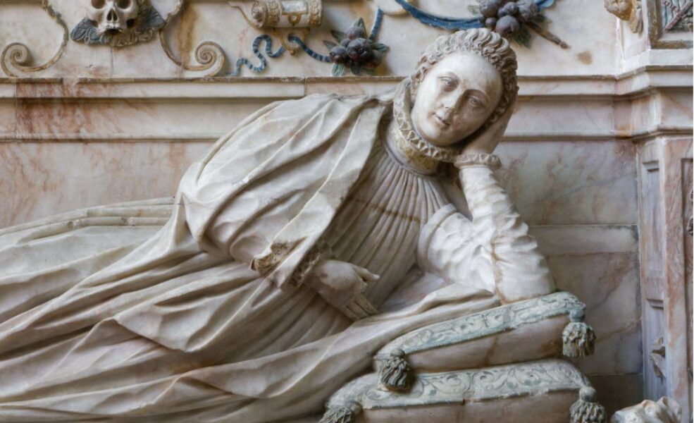 Tomb carving. Young woman in early C17 costume reclining on cushions in niche with baroque decoration and a skull