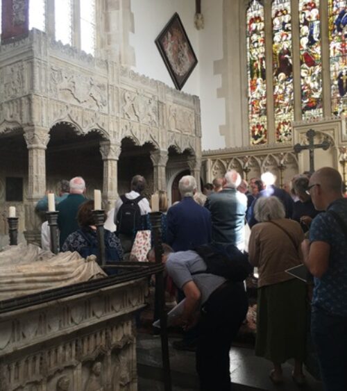 Group looking at tomb with elaborate canopy. Other monuments in background and foreground: funeral hatchment, stained glass.